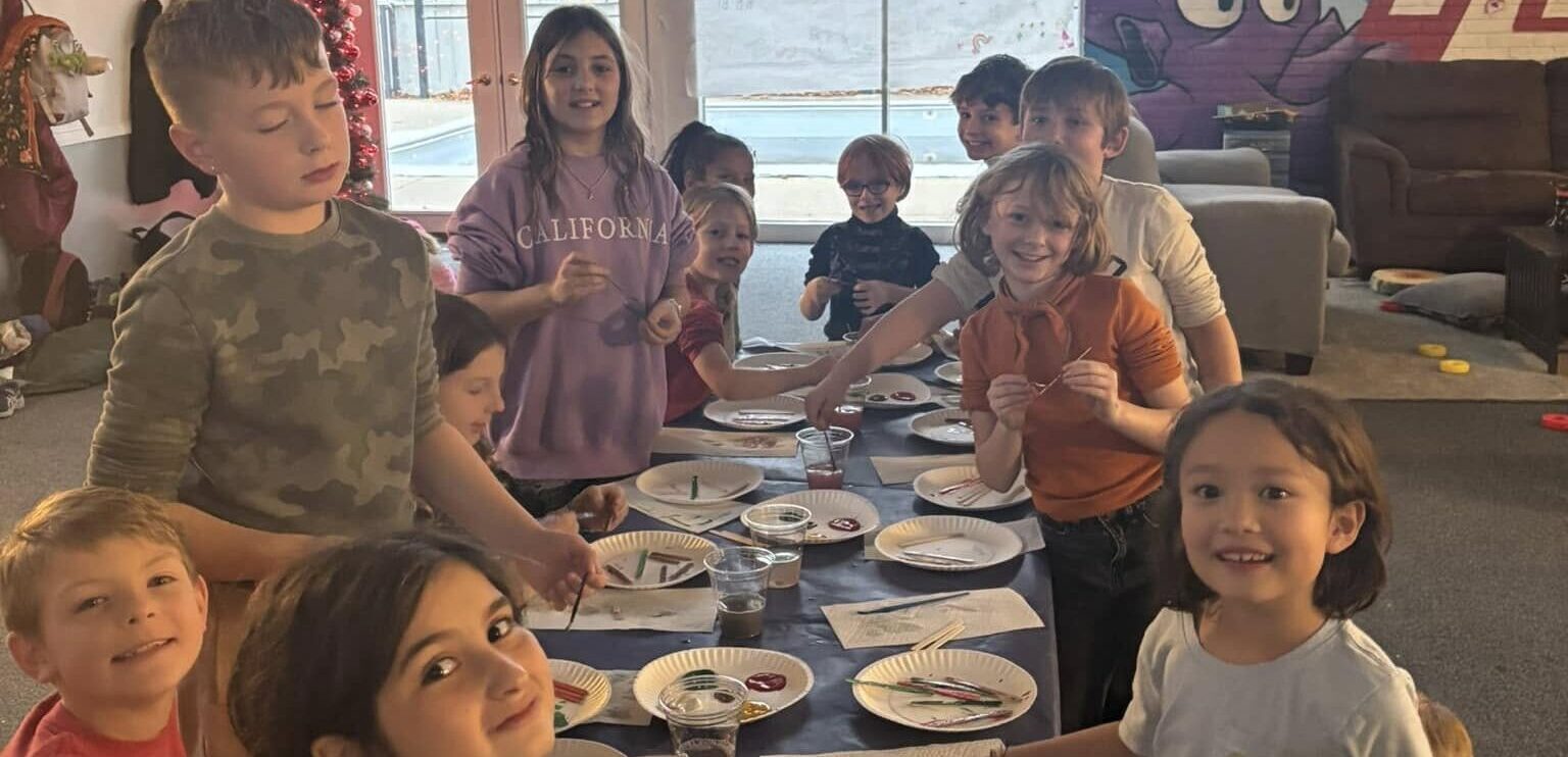 A group of elementary-age kids around a table indoors working on an art project in which multiple colors in abstract shapes are present on white paper plates. Some kids are holding brushes. A window to the back deck is in the background.