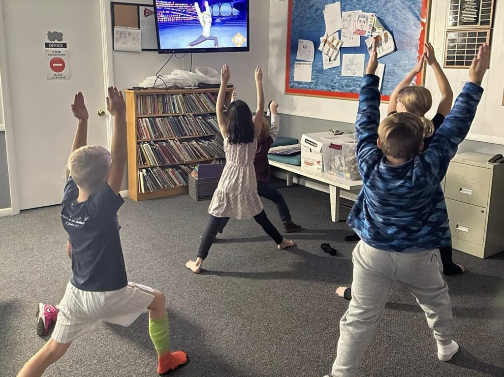 A circle of five elementary-aged children with their backs to the camera, arms stretched in the air, legs spread wide, mimicking a yoga or stretching pose from someone on a TV mounted in the upper corner of an indoor room with white rafters and a gray carpet.