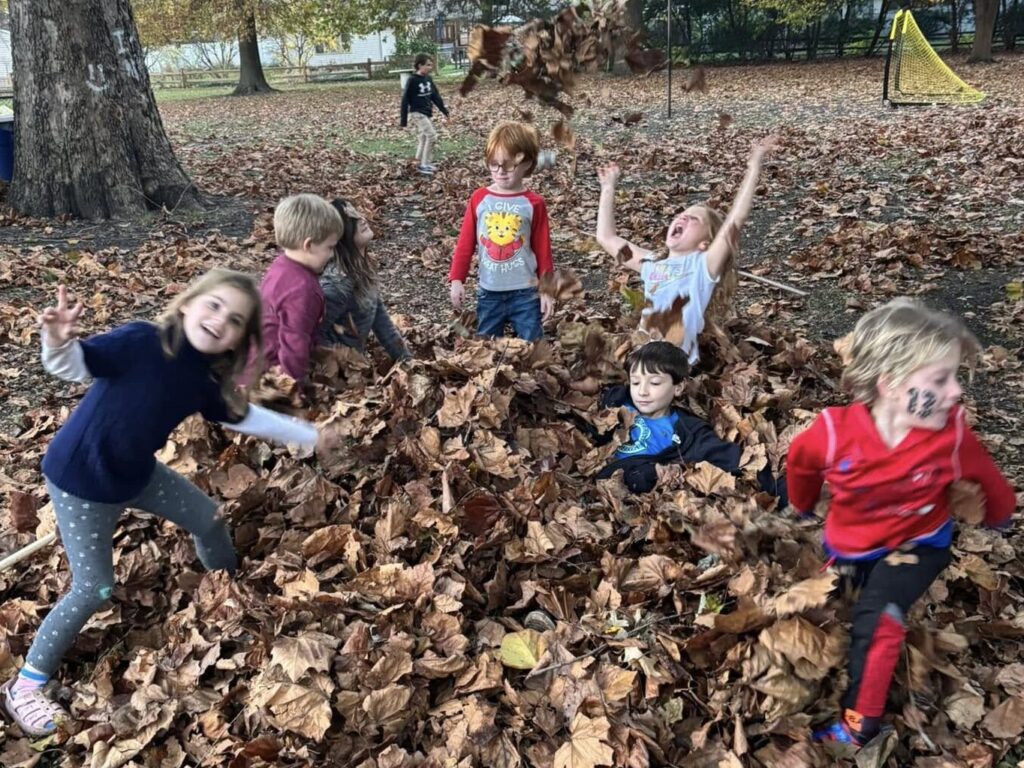 A semi-circle of five elementary-aged childen in a pile of brown leaves. A couple are looking at the camera and a couple are not. One is sitting in the leave pile up to his shoulders. Another is throwing leaves high into the air with her arms outstretched.