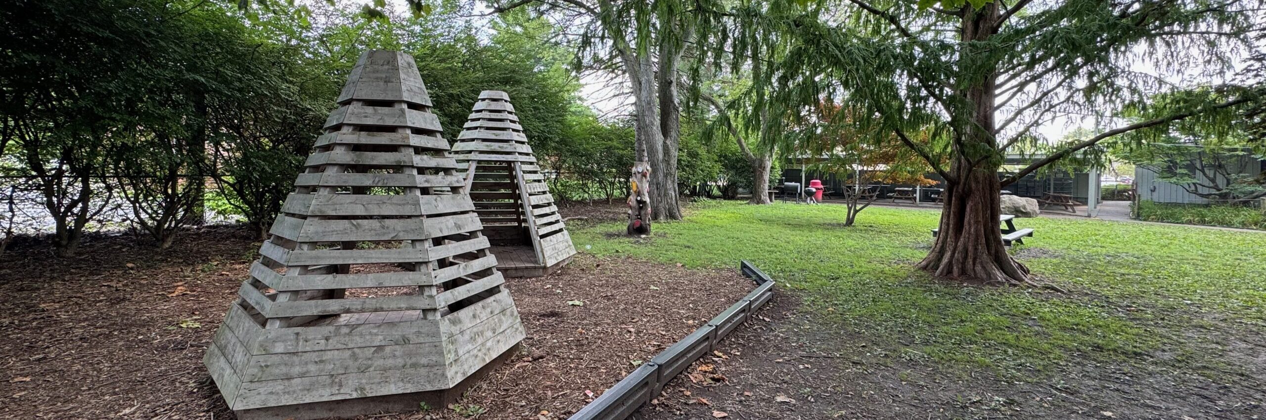 Two brown pyramid wooden structures about 1.5 meters high in a woodchip area like a playground. Grene grass, trees, and a building beyond.